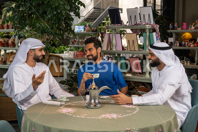 تناول المشروبات الساخنة، النقاش والحوار في الأجواء الهادئة، بناء علاقات اجتماعية ناجحة بين الأصحاب، قضاء وقت ممتع ومسلي مع الأصدقاء، صورة مقربة لرجلين عربيين خليجيين اماراتيين يرتديان الكندورة والغترة البيضاء يجلسان مع صديقهم في المقهي بايماءات السعادة والسرور
