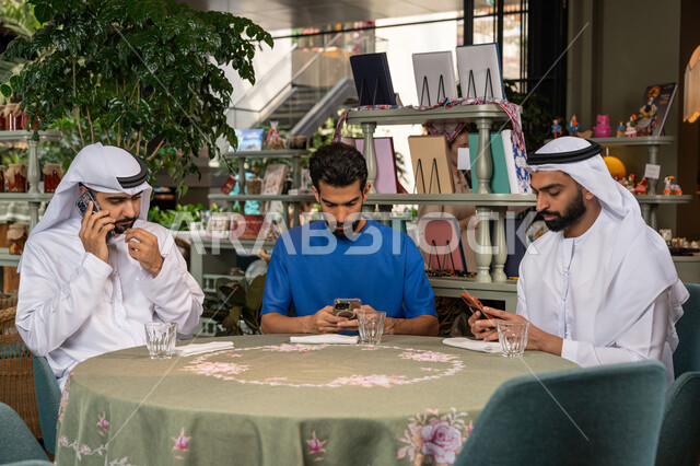اجراء مكالمة هاتفية، استخدام جهاز الهاتف المحمول، قضاء وقت ممتع مع الأصدقاء، التسلية والترفيه في العطلة، رجلان عربيان خليجان اماراتيان يرتديان الكندورة والغترة البيضاء يجلسان مع صديقهم في المقهى بايماءات السعادة والسرور