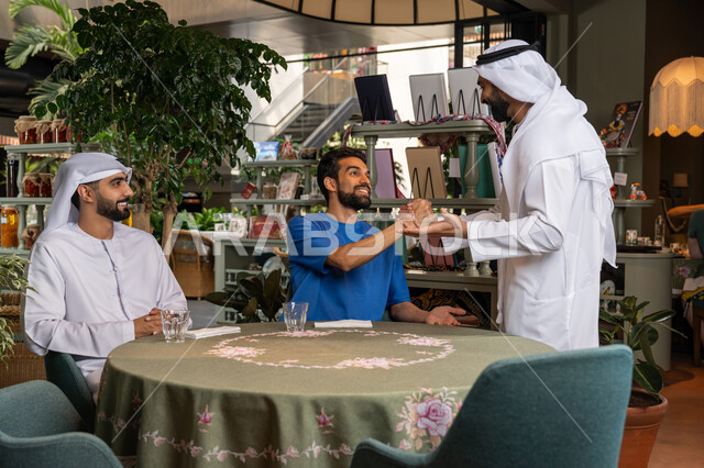 قضاء وقت ممتع ومسلي مع الأصدقاء في الخارج، الكافيهات والمطاعم في الامارات العربية المتحدة، التسلية والترفيه في العطلة، رجل عربي خليجي اماراتي يرتدي الكندورة والغترة البيضاء يقوم بالقاء التحية والسلام على اصدقائه في المقهى