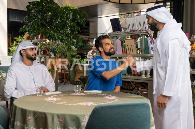 الكافيهات والمطاعم في الامارات العربية المتحدة، التسلية والترفيه في العطلة، رجل عربي خليجي اماراتي يرتدي الكندورة والغترة البيضاء يقوم بالقاء التحية والسلام على اصدقائه في المقهى، قضاء وقت ممتع ومسلي مع الأصدقاء في الخارج