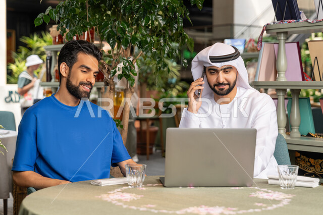 Gestures of happiness and pleasure, making a phone call via mobile, spending quality time with friends, close-up of a young Emirati Gulf Arab man wearing a kandura and ghutra sitting with his friend in a café, discussion and dialogue between colleagues, using a laptop to complete projects and tasks in the café