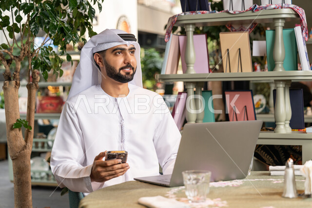 ادارة شؤون الاعمال وتنظيمها، استخدام الأجهزة التقنية الحديثة والمتطورة، تصفح مواقع التواصل الإجتماعي والعمل عن بعد في الكافيه، صورة مقربة لرجل عربي خليجي اماراتي مبتسم يرتدي الكندورة والغترة البيضاء يجلس  في المقهى ويستخدم الهاتف والحاسوب المحمول