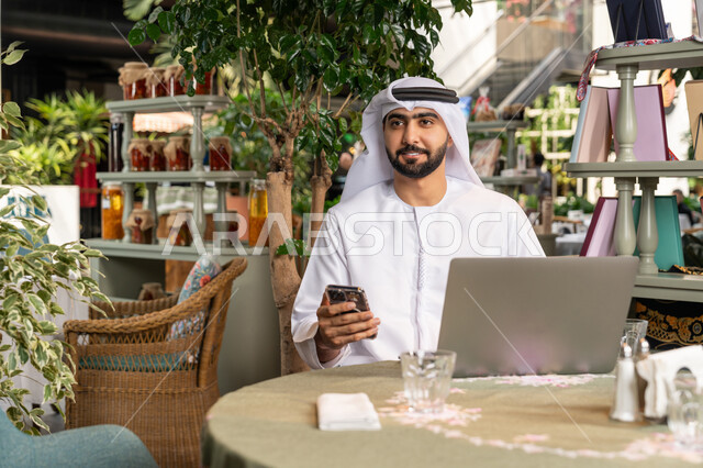 تصفح مواقع التواصل الإجتماعي والعمل عن بعد، استخدام الأجهزة التقنية الحديثة والمتطورة لانجاز المشاريع في الكافيه، صورة مقربة لرجل عربي خليجي اماراتي مبتسم يرتدي الكندورة والغترة البيضاء يجلس في المقهى ويستخدم الهاتف والحاسوب المحمول