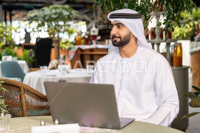 Using modern and advanced technical devices to complete projects, browsing social networking sites and working remotely, a close-up photo of a smiling Emirati Gulf Arab man wearing a white kandura and ghutra sitting on a comfortable chair in an upscale cafe and using a laptop with gestures of pleasure