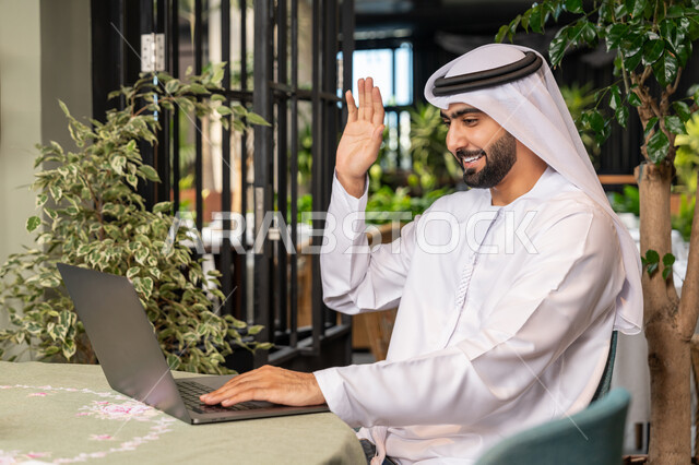 Making a video call via modern technical devices, using a laptop to complete projects in a café, a close-up of a smiling Emirati Gulf Arab man wearing a white kandura and ghutra sitting on a comfortable chair in a café and raising his hand in greeting gestures, working remotely