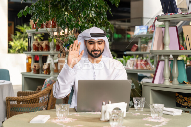 Working remotely, making a video call via modern technical devices, using a laptop to complete projects in a café, a close-up of a smiling Emirati Gulf Arab man wearing a white kandura and ghutra sitting on a comfortable chair in a café and raising his hand in greeting gestures