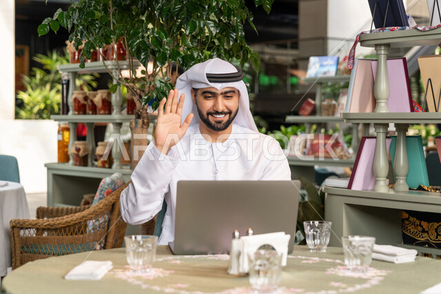 اجراء مكالمة فيديو عبر الاجهزة التقنية الحديثة، استخدام جهاز الحاسوب المحمول لانجاز المشاريع في المقهى، العمل عن بعد، صورة مقربة لرجل عربي خليجي اماراتي مبتسم يرتدي الكندورة والغترة البيضاء يجلس في الكافيه ويرفع يده بايماءات القاء التحية والسلام