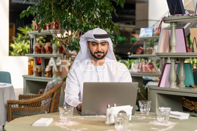 استخدام الأجهزة التقنية الحديثة والمتطورة لانجاز المشاريع في الكافيه، تصفح مواقع التواصل الإجتماعي والعمل عن بعد، صورة مقربة لرجل عربي خليجي اماراتي مبتسم يرتدي الكندورة والغترة البيضاء يجلس في المقهى ويستخدم جهاز الحاسوب المحمول بايماءات السرور