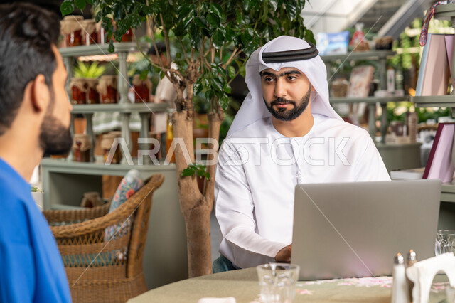 الحوار والنقاش بين زملاء العمل، استخدم جهاز الحاسوب المحمول لانجاز المشاريع والمهام في الكافيه، قضاء وقت ممتع مع الأصدقاء, صورة مقربة لشاب عربي خليجي إماراتي يرتدي الكندورة والغترة البيضاء يجلس مع صديقه في المقهى