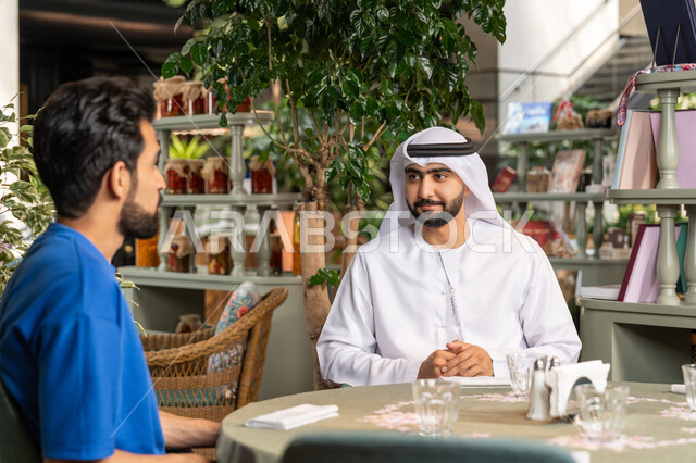 التسلية والترفيه في العطلة،  تبادل الاحاديث بين الأصدقاء، قضاء وقت ممتع في احدى الكافيهات، صورة مقربة لرجل عربي خليجي اماراتي يرتدي الكندورة والغترة البيضاء يجلس مع صديقه في المقهى