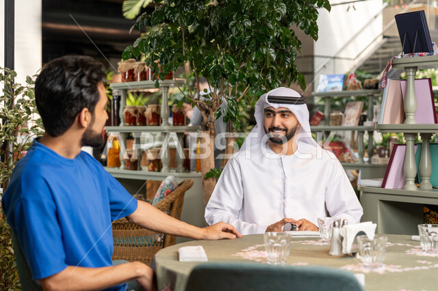 تبادل الاحاديث بين الأصدقاء، قضاء وقت ممتع في احدى الكافيهات، التسلية والترفيه في العطلة،  صورة مقربة لرجل عربي خليجي اماراتي يرتدي الكندورة والغترة البيضاء يجلس مع صديقه في المقهى