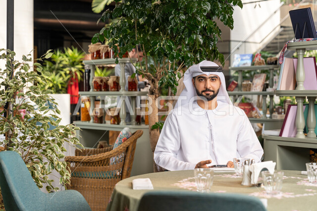 Having a fun and entertaining time in a café, looking at the camera with gestures of happiness and pleasure, a close-up of a smiling Emirati Gulf Arab man wearing a white kandura and ghutra sitting in a café holding a mobile phone, using a modern and advanced technical device