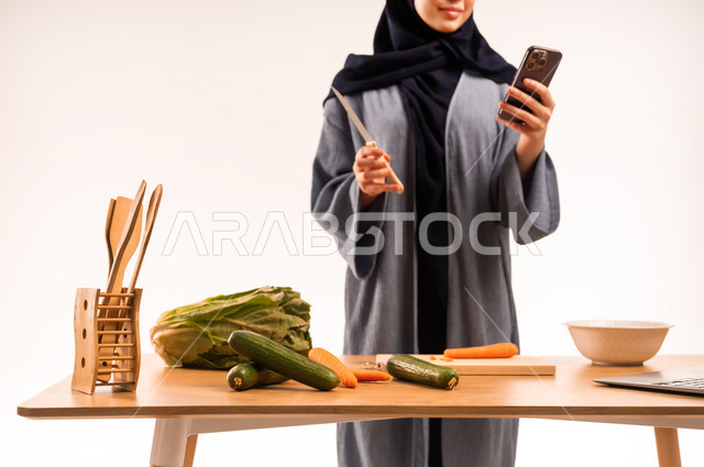 اعداد السلطة والأطباق الصحية، مشاركة تحضير الوصفات على مواقع التواصل الاجتماعي، مفهوم النظام الغذائي، بورتريه لامرأة عربية خليجية سعودية محجبة تقوم بإجراء بتدوين الملاحظات باستخدام الهاتف المحمول، مجموعة من الخضراوات الطازجة، خلفية بيضاء