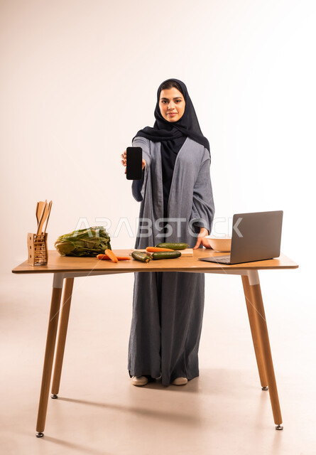 عرض شاشة سوداء فارغة، مشاركة تحضير الوصفات على مواقع التواصل الاجتماعي، إعداد السلطة والأطباق الصحية، مفهوم النظام الغذائي، استخدام الحاسوب المحمول، بورتريه لامرأة عربية خليجية سعودية محجبة تحمل بيدها الهاتف المحمول، مجموعة من الخضراوات الطازجة، صورة لكامل الجسم، خلفية بيضاء