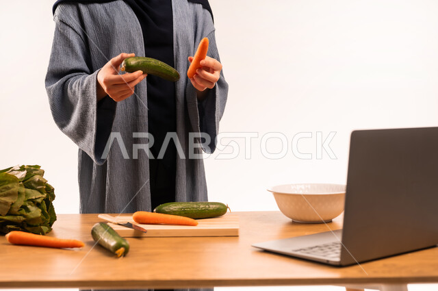 إعداد الأطباق الصحية، مفهوم النظام الغذائي، استخدام الحاسوب المحمول، بورتريه مقرب لامرأة عربية خليجية سعودية محجبة تقوم بصنع سلطة، مجموعة من الخضراوات الطازجة، مشاركة تحضير الوصفات على مواقع التواصل الاجتماعي، خلفية بيضاء