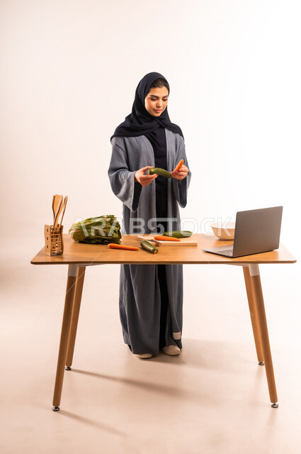إعداد الأطباق الصحية، مفهوم النظام الغذائي، استخدام الحاسوب المحمول، بورتريه لامرأة عربية خليجية سعودية محجبة تقوم بصنع سلطة، مجموعة من الخضراوات الطازجة، مشاركة تحضير الوصفات على مواقع التواصل الاجتماعي، صورة لكامل الجسم، خلفية بيضاء