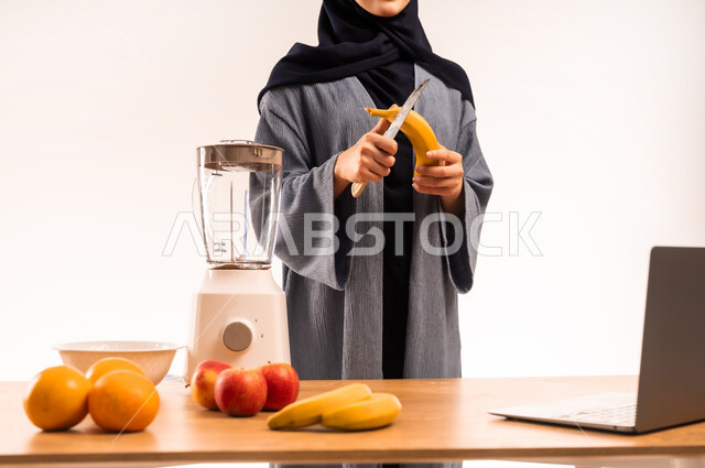 مفهوم النظام الغذائي، اعداد المشروبات الصحية، استخدام الحاسوب المحمول، بورتريه مقرب لامرأة عربية خليجية سعودية محجبة تقوم بصنع العصير باستخدام الخلاط، مجموعة من مكونات الفواكه الطازجة على الطاولة، مشاركة الوصفة عبر مواقع التواصل الاجتماعي، خلفية بيضاء