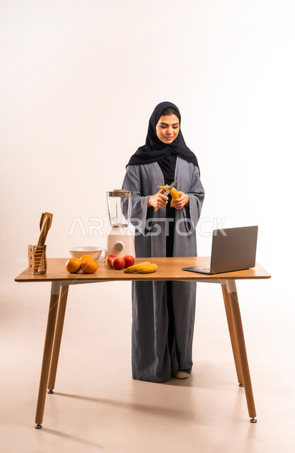 اعداد المشروبات الصحية، استخدام الحاسوب المحمول، بورتريه لامرأة عربية خليجية سعودية محجبة تقوم بصنع العصير باستخدام الخلاط، مجموعة من مكونات الفواكه الطازجة على الطاولة، مشاركة الوصفة عبر مواقع التواصل الاجتماعي، مفهوم النظام الغذائي، صورة لكامل الجسم، خلفية بيضاء
