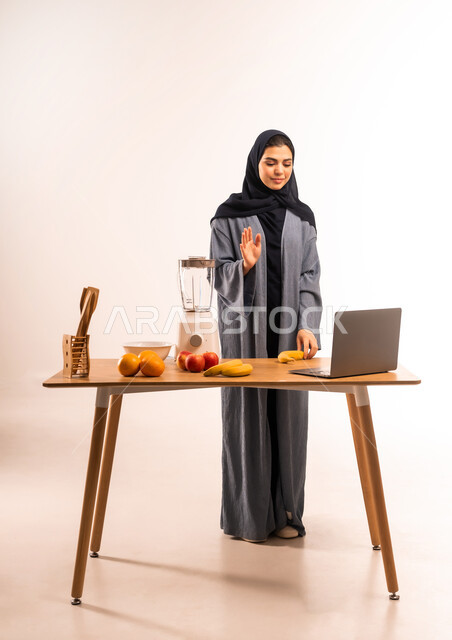 مفهوم النظام الغذائي، اعداد المشروبات الصحية، استخدام الحاسوب المحمول، بورتريه لامرأة عربية خليجية سعودية محجبة ترفع يدها بايماءات التحية والسلام، مجموعة من مكونات الفواكه الطازجة على الطاولة، مشاركة الوصفة عبر مواقع التواصل الاجتماعي، صورة لكامل الجسم، خلفية بيضاء