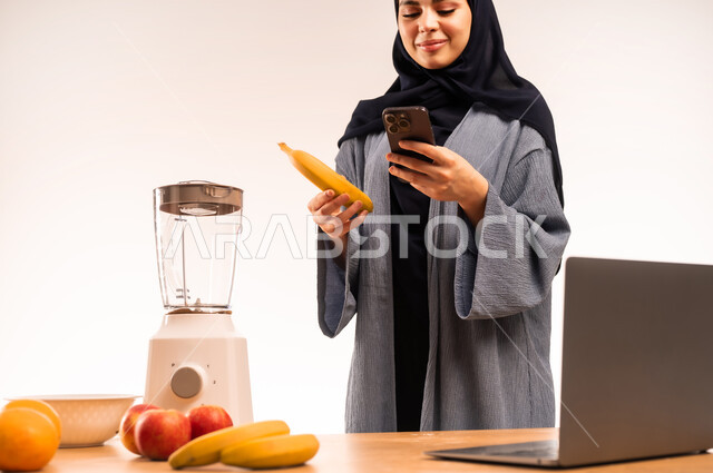 استخدام الحاسوب المحمول، مجموعة من الفواكه الطازجة على الطاولة، مشاركة الوصفة عبر مواقع التواصل الاجتماعي، مفهوم النظام الغذائي، اعداد المشروبات الصحية، بورتريه مقرب لامرأة عربية خليجية سعودية محجبة تحمل بيدها الموبايل تقوم بقراءة التعليمات، خلفية بيضاء