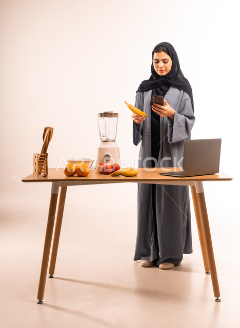 مجموعة من الفواكه الطازجة على الطاولة، مشاركة الوصفة عبر مواقع التواصل الاجتماعي، مفهوم النظام الغذائي، اعداد المشروبات الصحية، استخدام الحاسوب المحمول، بورتريه لامرأة عربية خليجية سعودية محجبة تحمل بيدها الموبايل تقوم بقراءة التعليمات، صورة لكامل الجسم، خلفية بيضاء