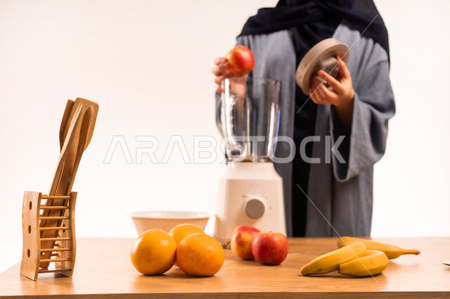 مفهوم النظام الغذائي، اعداد المشروبات الصحية، بورتريه مقرب لامرأة عربية خليجية سعودية محجبة تقوم بصنع العصير باستخدام الخلاط، مجموعة من مكونات الفواكه الطازجة على الطاولة، جهاز كهربائي منزلي، خلفية بيضاء