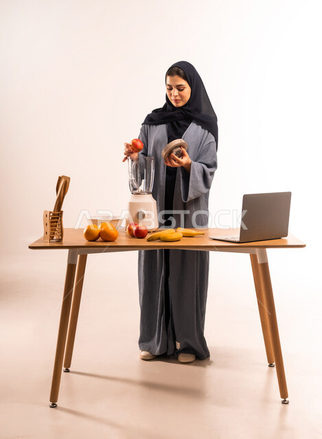 اعداد المشروبات الصحية، استخدام الحاسوب المحمول، بورتريه لامرأة عربية خليجية سعودية محجبة تقوم بصنع العصير باستخدام الخلاط، مجموعة من مكونات الفواكه الطازجة على الطاولة، مشاركة الوصفة عبر مواقع التواصل الاجتماعي، مفهوم النظام الغذائي، صورة لكامل الجسم، خلفية بيضاء