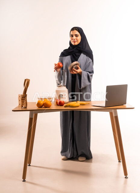 استخدام الحاسوب المحمول، بورتريه لامرأة عربية خليجية سعودية محجبة تقوم بصنع العصير باستخدام الخلاط، مجموعة من مكونات الفواكه الطازجة على الطاولة، مشاركة الوصفة عبر مواقع التواصل الاجتماعي، إعداد المشروبات الصحية، مفهوم النظام الغذائي، صورة لكامل الجسم، خلفية بيضاء