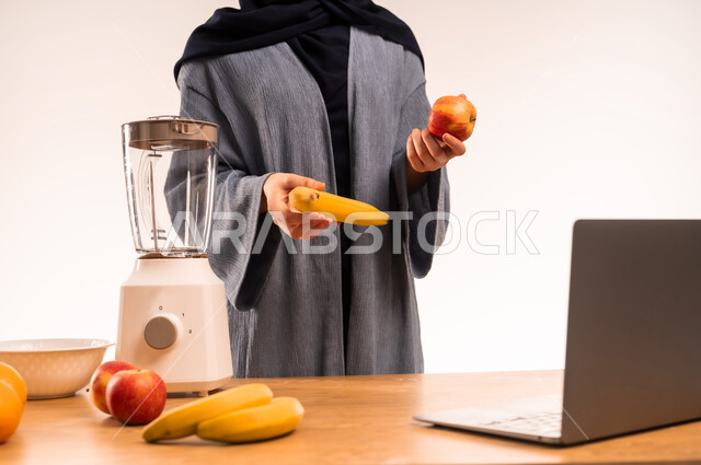 مفهوم النظام الغذائي، اعداد المشروبات الصحية، استخدام الحاسوب المحمول، بورتريه مقرب لامرأة عربية خليجية سعودية محجبة تقوم بصنع العصير باستخدام الخلاط، مجموعة من مكونات الفواكه الطازجة على الطاولة، مشاركة الوصفة عبر مواقع التواصل الاجتماعي، خلفية بيضاء