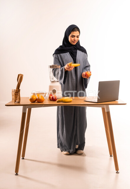 استخدام الحاسوب المحمول، بورتريه لامرأة عربية خليجية سعودية محجبة تقوم بصنع العصير باستخدام الخلاط، مجموعة من مكونات الفواكه الطازجة على الطاولة، مشاركة الوصفة عبر مواقع التواصل الاجتماعي، إعداد المشروبات الصحية، مفهوم النظام الغذائي، صورة لكامل الجسم، خلفية بيضاء