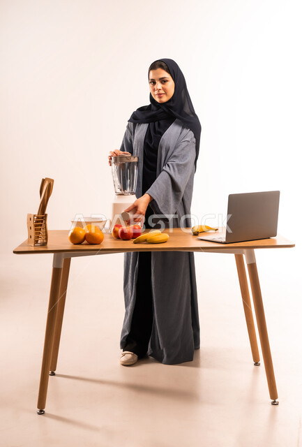 اعداد المشروبات الصحية، استخدام الحاسوب المحمول، بورتريه لامرأة عربية خليجية سعودية محجبة تقوم بصنع العصير باستخدام الخلاط، مجموعة من مكونات الفواكه الطازجة على الطاولة، مشاركة الوصفة عبر مواقع التواصل الاجتماعي، مفهوم النظام الغذائي، صورة لكامل الجسم، خلفية بيضاء