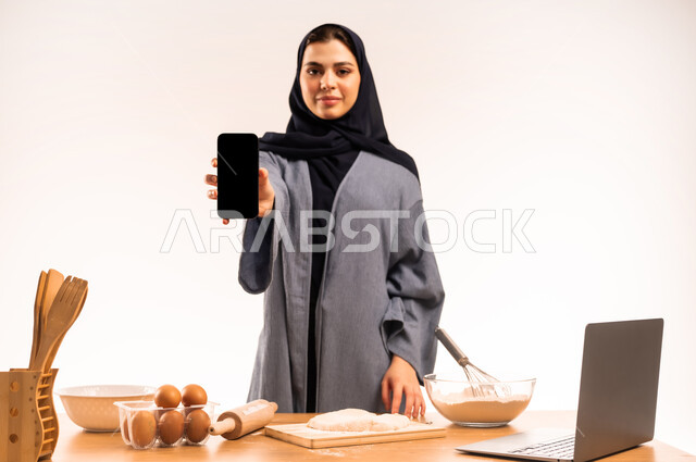 مسؤولية ربة المنزل، مشاركة الوصفة عبر مواقع التواصل الاجتماعي، استخدام الحاسوب المحمول، أدوات خاصة بالمطبخ والعجين، إعداد المخبوزات والمعجنات، بورتريه مقرب لامرأة عربية خليجية سعودية محجبة تحمل بيدها الهاتف المحمول تقوم بعرض شاشة سوداء فارغة، خلفية بيضاء