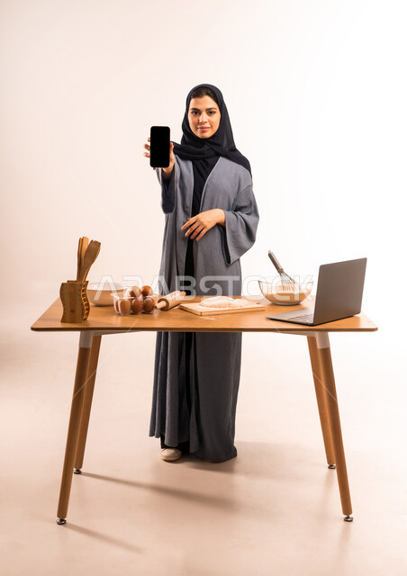 مشاركة الوصفة عبر مواقع التواصل الاجتماعي، استخدام الحاسوب المحمول، أدوات خاصة بالمطبخ والعجين، إعداد المخبوزات والمعجنات، بورتريه لامرأة عربية خليجية سعودية محجبة تحمل بيدها الهاتف المحمول تقوم بعرض شاشة سوداء فارغة، صورة لكامل الجسم، خلفية بيضاء