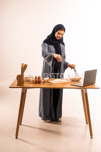Using laptop, kitchen and dough tools, preparing baked goods and pastries, sharing recipe via social media, housewife responsibility, portrait of a veiled Saudi Arabian Gulf woman holding a hand mixer and sprinkling flour, full body image, white background