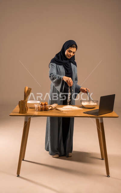 مشاركة الوصفة عبر مواقع التواصل الاجتماعي، استخدام الحاسوب المحمول، أدوات خاصة بالمطبخ والعجين، إعداد المخبوزات والمعجنات، مسؤولية ربة المنزل، بورتريه لامرأة عربية خليجية سعودية محجبة تحمل الخفاقة اليدوية تقوم برش الطحين، صورة لكامل الجسم، خلفية بنية