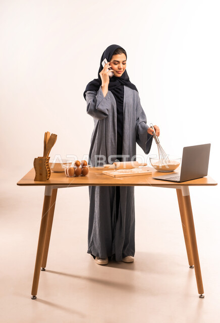 إعداد المخبوزات والمعجنات، مشاركة الوصفة عبر مواقع التواصل الاجتماعي، استخدام الحاسوب المحمول، مسؤولية ربة المنزل، بورتريه لامرأة عربية خليجية سعودية محجبة تحمل الخفاقة اليدوية تقوم باجراء مكالمة عبر الموبايل، أدوات خاصة بالمطبخ والعجين، الجسم الكامل، خلفية بيضاء
