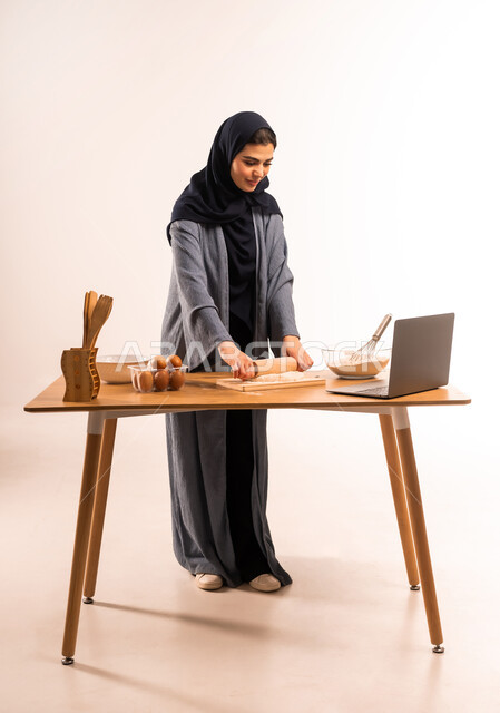 تحضير المكونات لصناعة الخبز، استخدام الحاسوب المحمول، بورتريه لامرأة عربية خليجية سعودية محجبة تحمل بيدها الشوبك (النشابة) تقوم بمشاركة وصفة العجين على مواقع التواصل الاجتماعي، أدوات خاصة بالمطبخ، إعداد المخبوزات والمعجنات الشهية، صورة لكامل الجسم، خلفية بيضاء