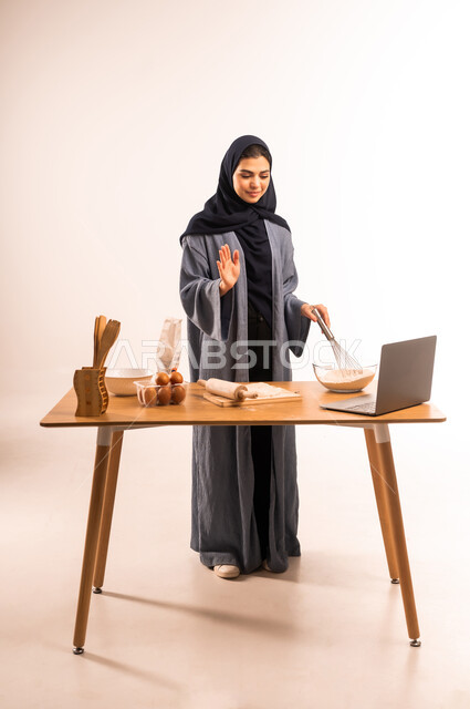 Sharing dough recipe on social media, using laptop, portrait of a veiled Saudi Arabian Gulf woman holding a hand mixer raising her hand in greeting gestures, kitchen tools, preparing delicious baked goods and pastries, full body photo, white background