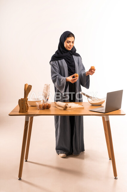 إعداد المخبوزات والمعجنات الشهية، تحضير المكونات لصناعة الخبز، استخدام الحاسوب المحمول، بورتريه لامرأة عربية خليجية سعودية محجبة تحمل بيدها البيض تقوم بمشاركة وصفة العجين على مواقع التواصل الاجتماعي، أدوات خاصة بالمطبخ، صورة لكامل الجسم، خلفية بيضاء