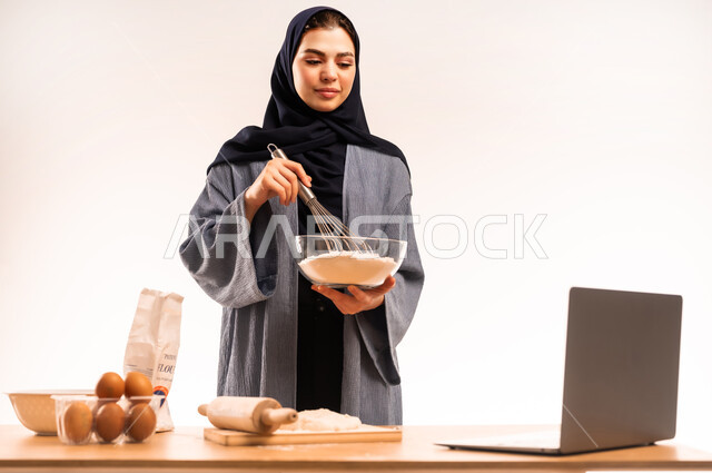 Using laptop, preparing ingredients for bread making, close-up portrait of veiled Saudi Arabian Gulf woman holding hand mixer sharing dough recipe on social media, kitchen tools, preparing delicious baked goods and pastries, white background