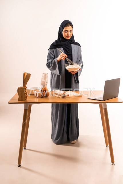Preparing ingredients for making bread, using laptop, portrait of a veiled Saudi Arabian Gulf woman holding a hand mixer sharing dough recipe on social media, kitchen tools, preparing delicious baked goods and pastries, full body image, white background