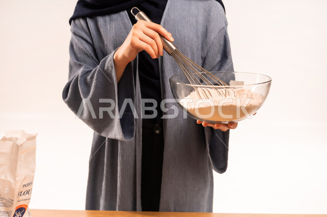 إعداد المخبوزات والمعجنات الشهية، مسؤولية ربة المنزل، تجهيز المكونات لصناعة الخبز، بورتريه مقرب لايدي امرأة عربية خليجية سعودية محجبة تحمل الخفاقة اليدوية تقوم بتحضير العجين، أدوات خاصة بالمطبخ، خلفية بيضاء