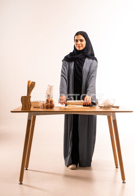 استخدام أداة فرد العجين، بورتريه لامرأة عربية خليجية سعودية محجبة تحمل بيدها الشوبك (النشابة) تقوم بتحضير الطعام، تجهيز المكونات لصناعة الخبز، أدوات خاصة بالمطبخ، إعداد المخبوزات والمعجنات الشهية، مسؤولية ربة المنزل، صورة لكامل الجسم، خلفية بيضاء
