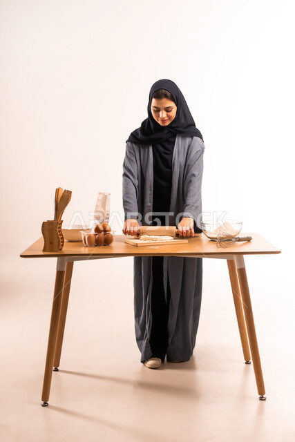 تجهيز المكونات لصناعة الخبز، استخدام أداة فرد العجين، بورتريه لامرأة عربية خليجية سعودية محجبة تحمل بيدها الشوبك (النشابة) تقوم بتحضير الطعام، أدوات خاصة بالمطبخ، إعداد المخبوزات والمعجنات الشهية، مسؤولية ربة المنزل، صورة لكامل الجسم، خلفية بيضاء