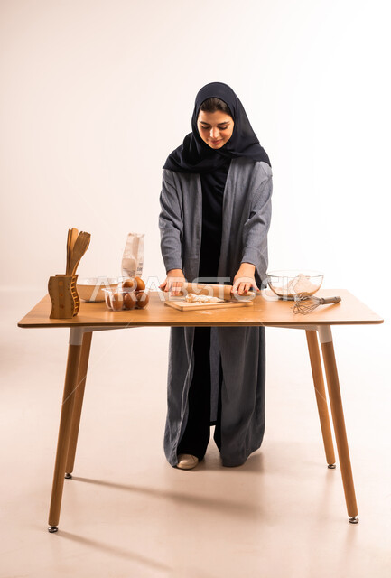 Preparing ingredients for making bread, using a dough roller, portrait of a veiled Saudi Arabian Gulf woman holding a rolling pin in her hand preparing food, kitchen tools, preparing delicious baked goods and pastries, housewife responsibility, full body image, white background