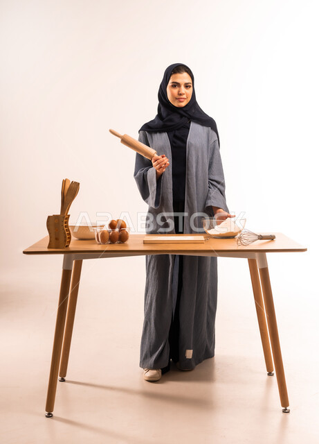 استخدام أداة فرد العجين، بورتريه لامرأة عربية خليجية سعودية محجبة تحمل بيدها الشوبك (النشابة) تقوم بتحضير الطعام، تجهيز المكونات لصناعة الخبز، أدوات خاصة بالمطبخ، إعداد المخبوزات والمعجنات الشهية، مسؤولية ربة المنزل، صورة لكامل الجسم، خلفية بيضاء