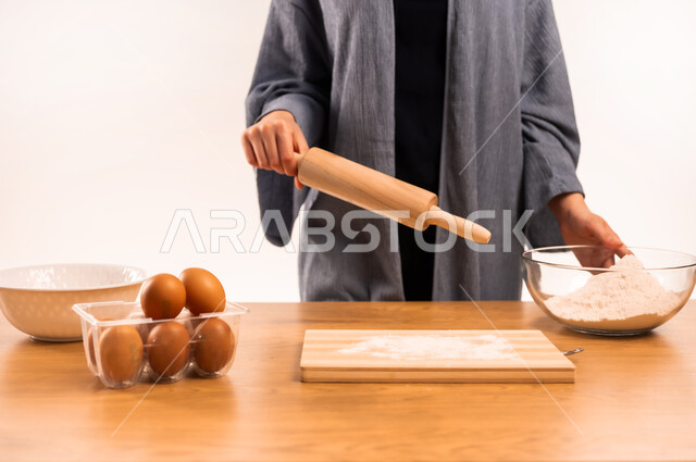 إعداد المخبوزات والمعجنات الشهية، تجهيز المكونات لصناعة الخبز، استخدام أداة فرد العجين، بورتريه مقرب لايدي امرأة عربية خليجية سعودية محجبة تحمل بيدها الشوبك (النشابة) تقوم بتحضير الطعام، أدوات خاصة بالمطبخ، مسؤولية ربة المنزل، خلفية بيضاء