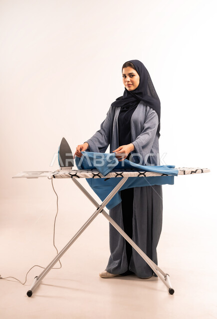 تجفيف وتنظيم الملابس، انجاز المهام والأعمال الروتينية، بورتريه لامرأة عربية خليجية سعودية محجبة ترتدي العباءة تقوم بكي الملابس باستخدام المكواة، استخدام الجهاز الكهربائي، مفهوم التدبير المنزلي، صورة لكامل الجسم، خلفية بيضاء