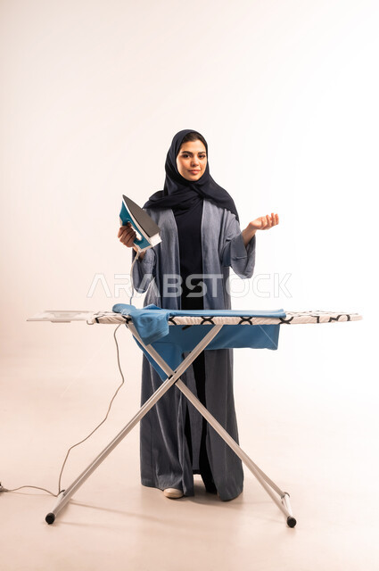 انجاز المهام والأعمال الروتينية، بورتريه لامرأة عربية خليجية سعودية محجبة ترتدي العباءة تقوم بكي الملابس باستخدام المكواة، استخدام الجهاز الكهربائي، مفهوم التدبير المنزلي، تجفيف وتنظيم الملابس، صورة لكامل الجسم، خلفية بيضاء