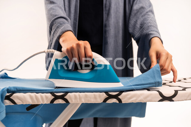 Using electrical appliance, drying and organizing clothes, completing chores and routines, close-up portrait of veiled Saudi Arabian Gulf woman wearing abaya ironing clothes with iron, housekeeping concept, white background